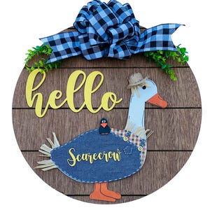 May include: A round wooden door hanger with a brown woodgrain finish. The door hanger features a white goose with a straw hat and a blue denim scarecrow costume. The goose is holding a small black bird on its back. The door hanger also features a yellow "hello" sign and a blue and black checkered bow.