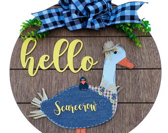 May include: A round wooden door hanger with a brown woodgrain finish. The door hanger features a white goose with a straw hat and a blue denim scarecrow costume. The goose is holding a small black bird on its back. The door hanger also features a yellow "hello" sign and a blue and black checkered bow.