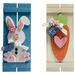 May include: Two wooden Easter decorations. The first is a blue and white bunny with a pink nose and a pink flower on its head. The second is a white wooden board with a carrot and a pink heart. The carrot has a green top and a tag that says "Carrots 25¢".