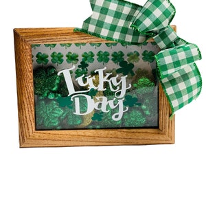 May include: A wooden frame with a green and gold glitter background and the words "Lucky Day" in white. The frame is decorated with a green and white checkered ribbon.