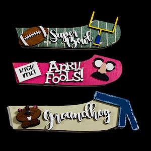 May include: Three wooden signs with different themes. The first sign is green and white with a football and the words "Super Bowl". The second sign is pink with a mustache, glasses, and the words "April Fools!" and "Kick me!" written on a note. The third sign is beige with a brown groundhog and the word "Groundhog" in white.