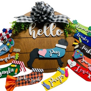 May include: A wooden door hanger with a black and white checkered bow, the word "hello" in white script, and a black pug silhouette wearing a blue and white vest with a pink heart.  The hanger is decorated with various colorful, festive, and seasonal words and phrases, including "Winter", "Turkey", "Summer", "Love XO", "Spring", "Pumpkin", "Santa", "Trick or Treat", "Peeps", "Be Mine", "Shinanigans", and "American".