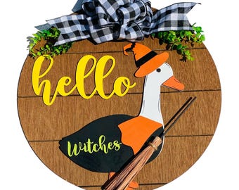 May include: A round wooden door hanger with a black and white checkered bow, green leaves, and a yellow "hello" sign. A cartoon goose wearing a witch's hat and holding a broom says "witches".