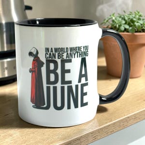 May include: White ceramic mug with a black interior and handle. The mug features the text "IN A WORLD WHERE YOU CAN BE ANYTHING BE A JUNE" with a red and black graphic of a person in a red cloak.