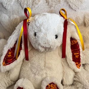 May include: A plush white bunny with red and gold accents. The inside of the ears and the soles of the feet feature a maroon fabric with a gold design. The bunny has red and gold ribbons tied to its ears.