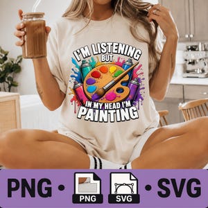 Könnte beinhalten: Ein beiges T-Shirt mit einer bunten Grafik aus einer Farbpalette, einem Pinsel und Sprühdosen. Der Text lautet I'M LISTENING BUT IN MY HEAD I'M PAINTING. Das Design enthält lebendige Farbspritzer in den Farbtönen Blau, Pink und Violett.