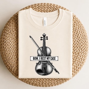 Könnte beinhalten: Ein cremefarbenes T-Shirt mit einer schwarz-weißen Grafik einer Geige und eines Bogens. Über die Mitte des Instruments verläuft ein rechteckiges Banner mit dem schwarzen Text "BOW, I REST MY CASE" in einer fetten, serifenlosen Schrift.