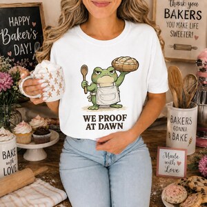 Könnte beinhalten: Ein weißes Kurzarm-T-Shirt mit einer Grafik eines grünen Frosches in einer weißen Schürze, der einen Holzlöffel und einen Laib Brot hält, mit dem Text "WE PROOF AT DAWN" in dunkelbraunen Buchstaben unter der Illustration.