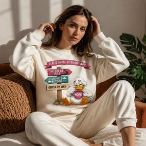 Pode incluir: Uma camisola de gola redonda cor de creme com o gráfico de um pato de desenho animado a segurar uma caneca cor-de-rosa com a palavra NOPE. Por cima do pato, há um poste de madeira com três sinais: PLACES I WON'T BE GOING TODAY, THE EXTRA MILE, ABOVE & BEYOND.