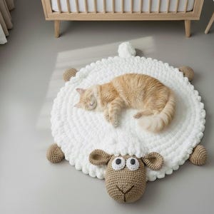 Op de afbeelding: Een witte, gehaakte schapenvormige vloerkleed met een slapende oranje kat. Het kleed heeft bruine gehaakte oren en poten en een gehaakt schapengezicht. De kat ligt opgerold op het kleed en ziet er comfortabel en ontspannen uit.