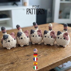 May include: A row of five crocheted cat figures. Each cat has a cream-colored body, with patches of pink and gray. The tails are striped. The word "PATTERN" is written in black capital letters above the cats.