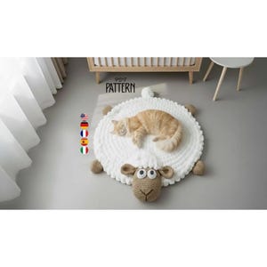 May include: A round white crocheted rug shaped like a sheep with a tan head, ears, and feet. The sheep has large white eyes with black pupils and a stitched nose. The text PDF PATTERN is visible above the sheep, along with five small circular flag icons.