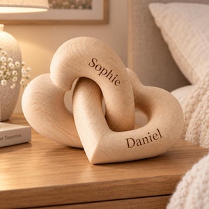 May include: A light-coloured wooden sculpture consisting of two interlocking heart shapes. The name Sophie is engraved on the upper heart, and the name Daniel is engraved on the lower heart. The wood features a visible grain pattern throughout the surface.