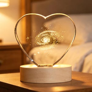 May include: A heart-shaped clear acrylic plaque featuring an etched golden spiral galaxy design with small stars. The plaque is mounted on a cylindrical light-colored wooden base that illuminates the acrylic from below with a warm glow.