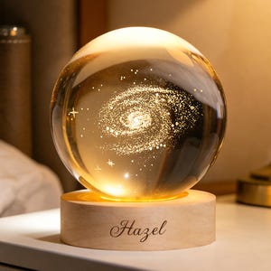 Könnte beinhalten: Eine klare Glaskugel mit einem innen laser-gravierten Spiralgalaxie-Design und kleinen Sternakzenten. Die Kugel ruht auf einem zylindrischen, hellen Holzsockel, auf dessen Vorderseite der Name Hazel in einer Schreibschrift eingraviert ist.