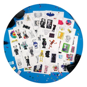 Op de afbeelding: Een verzameling kleurrijke stickers met verschillende stripfiguren en ontwerpen. De stickers zijn gerangschikt op witte vellen tegen een blauwe achtergrond, met een paar verspreide confetti. De ontwerpen omvatten figuren in verschillende poses en kleuren.