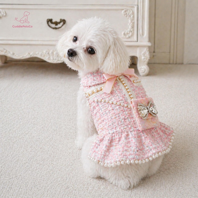 May include: A small white dog wearing a pink tweed dress with a bow and pearl detailing. The dress features a frilled skirt and a small pocket with a jewelled bow. The dress is from CuddlePetsCo.
