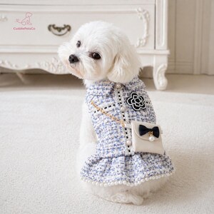 May include: A small white dog dressed in a blue and white tweed dress, featuring a black flower detail and a cream-coloured purse. The dress is adorned with pearl accents and a gold chain strap. The logo "CuddlePetsCo" is visible.