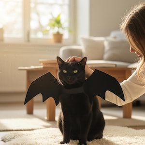 May include: A black cat wearing a set of black bat wings attached to a collar. The wings are made of a solid black material with a scalloped edge design, extending from the cat's shoulders. The cat has bright green eyes and sits upright.