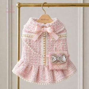 May include: A pink tweed dog dress with a matching bow and a small pocket with a jeweled bow. The dress features pearl accents and a ruffled skirt. The dress is hanging on a gold hanger.