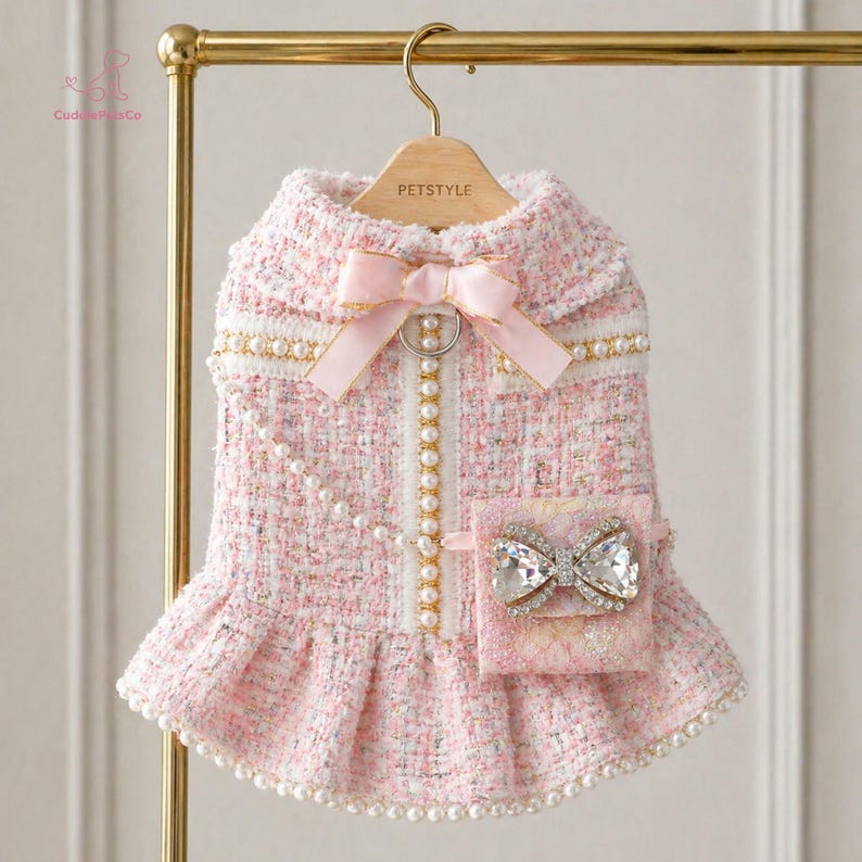 May include: A pink tweed dog dress with a matching bow and a small pocket with a jeweled bow. The dress features pearl accents and a ruffled skirt. The dress is hanging on a gold hanger.