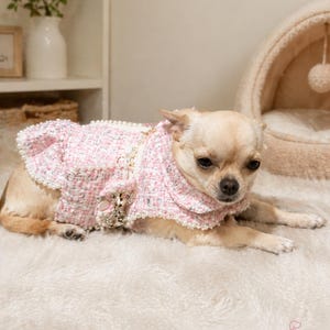 May include: A small dog wearing a pink tweed jacket with a decorative bow and pearl accents. The dog is lying on a fluffy, white surface. The jacket has a ruffled collar and trim. The CuddlePetsCo logo is visible in the bottom right corner.