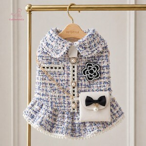 May include: A blue and white tweed dog dress with a white collar and pearl accents. The dress features a small pocket with a black bow and a decorative flower. A gold chain strap is attached to the dress.