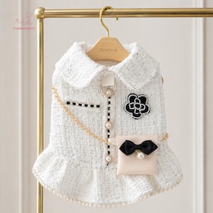 May include: A white tweed dog dress with a Peter Pan collar, black detailing, and a small purse with a black bow. It has a gold chain and pearl embellishments, hanging on a gold hanger.