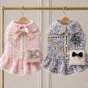 May include: Two dog dresses on a gold rail. The pink dress features a bow, jeweled pocket, and pearl detailing. The blue dress has a black bow, a flower, a pearl chain, and pearl accents. Both dresses have a frilled hem.