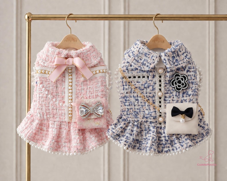 May include: Two dog dresses on a gold rail. The pink dress features a bow, jeweled pocket, and pearl detailing. The blue dress has a black bow, a flower, a pearl chain, and pearl accents. Both dresses have a frilled hem.