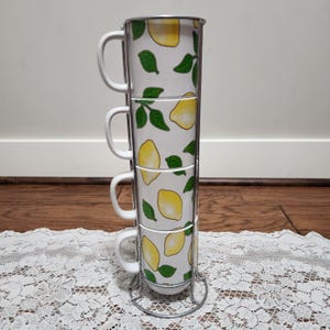 May include: A set of four white ceramic mugs stacked in a metal rack. Each mug is decorated with yellow lemons and green leaves. The mugs have white handles and are designed for serving beverages.