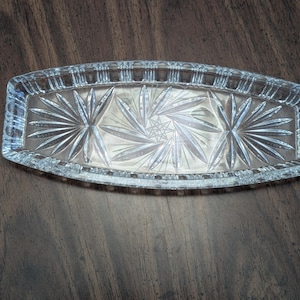 May include: A clear, rectangular glass serving dish with a starburst design in the center. The dish has a decorative, ridged border. The glass is transparent, allowing light to pass through, and is set against a dark wooden surface.