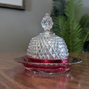 May include: A clear glass candy dish with a diamond pattern on the lid and a ruby red base. The dish is on a wooden table. The lid has a decorative finial.