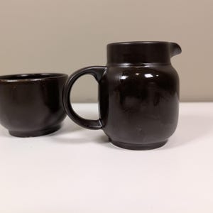May include: A dark brown ceramic creamer jug with a handle and a small bowl. The jug has a rounded body and a spout. The bowl is round with a slightly flared rim. Both items are on a white surface.