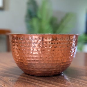 Peut inclure: Un bol rond en métal fabriqué en cuivre avec une texture martelée distincte sur toute sa surface extérieure. Le bol présente une finition métallique brun rougeâtre chaleureuse et un large sommet ouvert avec un bord arrondi et lisse.