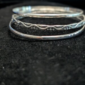 May include: A set of three silver bangles stacked together. The top and bottom bangles have a smooth, polished finish. The middle bangle features a textured design with a repeating wave pattern and small dots along the band's surface.