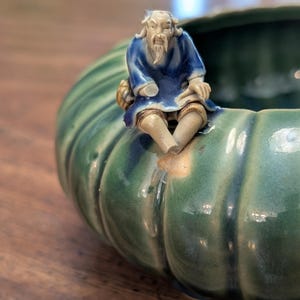 Puede incluir: Un cuenco de cerámica verde con exterior acanalado y una figura sentada de un anciano con túnicas azules. El cuenco tiene un acabado brillante y está sobre una superficie de madera. La figura está detallada con barba y pelo blancos.