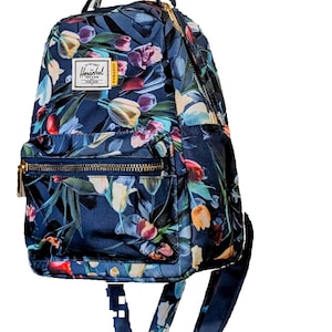 May include: A navy blue backpack with a floral tulip design in red, yellow, and purple. The bag has a front zipped pocket, gold-coloured zips, and the "Herschel" brand label.