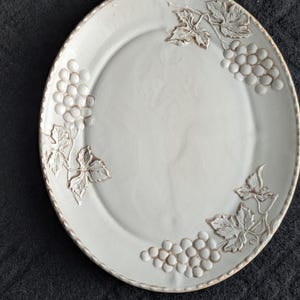 May include: Off-white ceramic plate featuring an embossed grape and leaf pattern. The plate has a raised rim and a textured finish. The grape clusters and leaves are a lighter shade, providing a subtle contrast. A decorative serving plate.
