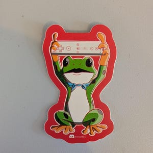 May include: A sticker depicting a cartoon frog with a moustache and a blue bow tie, holding a white game controller. The frog is green with orange hands and feet, set against a red background. The text "boxes1964" is at the bottom.