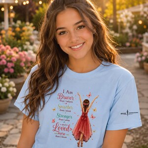 May include: A light blue t-shirt featuring a graphic of a person in a red dress with arms raised, surrounded by butterflies. The text reads: I Am Braver than I believe, Smarter than I think, Stronger than I seem and Loved more than I know.