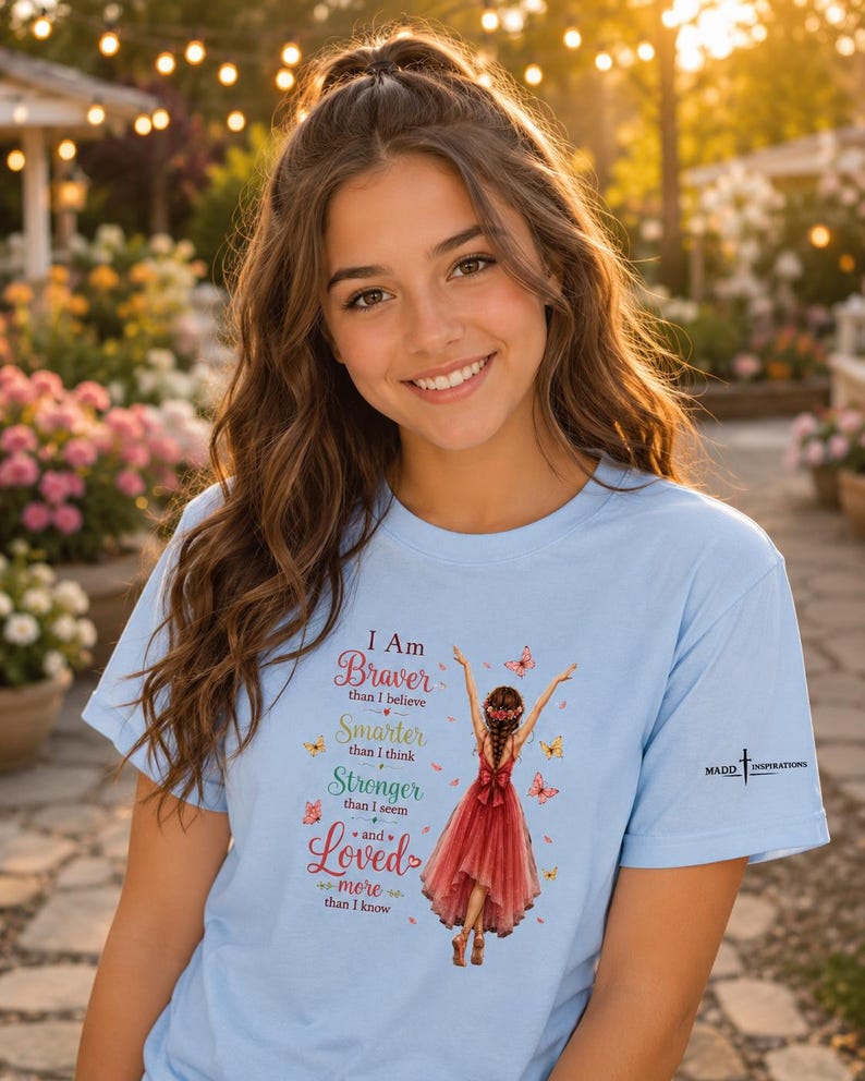 May include: A light blue t-shirt featuring a graphic of a person in a red dress with arms raised, surrounded by butterflies. The text reads: I Am Braver than I believe, Smarter than I think, Stronger than I seem and Loved more than I know.