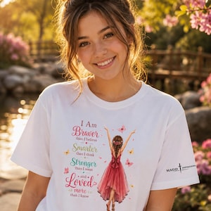 May include: A white t-shirt featuring a graphic of a person in a red dress with arms raised, surrounded by butterflies. The text reads: I Am Braver than I believe, Smarter than I think, Stronger than I seem and Loved more than I know. MADD INSPIRATIONS logo.