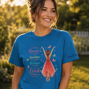 May include: A blue short-sleeved t-shirt featuring a graphic of a person in a red dress with butterflies. The text reads: I Am Braver than I believe, Smarter than I think, Stronger than I seem, and Loved more than I know. The shirt has a small logo on the sleeve.