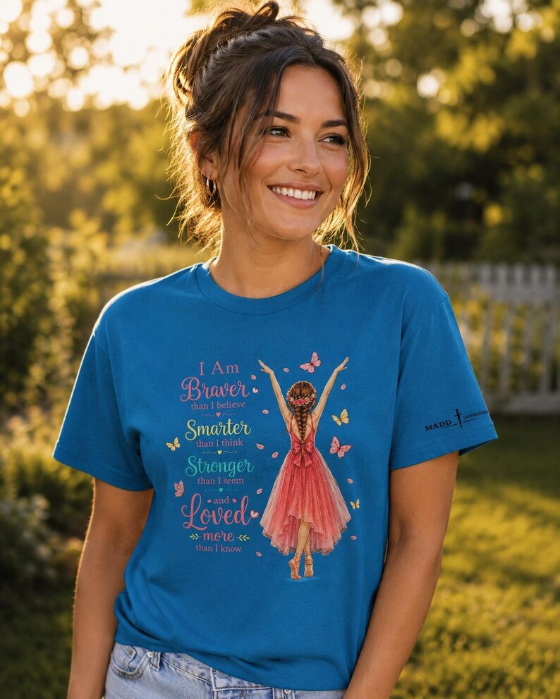 May include: A blue short-sleeved t-shirt featuring a graphic of a person in a red dress with butterflies. The text reads: I Am Braver than I believe, Smarter than I think, Stronger than I seem, and Loved more than I know. The shirt has a small logo on the sleeve.