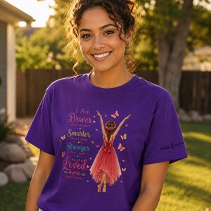 May include: A purple short-sleeved t-shirt featuring a graphic of a person in a red dress with arms raised, surrounded by butterflies. The text reads: I Am Braver than I believe, Smarter than I think, Stronger than I seem, and Loved more than I know.
