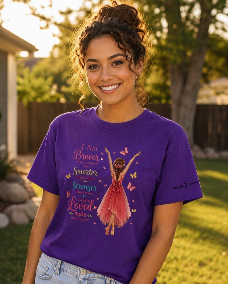 May include: A purple short-sleeved t-shirt featuring a graphic of a person in a red dress with arms raised, surrounded by butterflies. The text reads: I Am Braver than I believe, Smarter than I think, Stronger than I seem, and Loved more than I know.