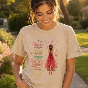 May include: A beige short-sleeved t-shirt featuring a graphic of a person in a red dress with arms raised, surrounded by butterflies. The text reads: I Am Braver than I believe, Smarter than I think, Stronger than I seem, and Loved more than I know.