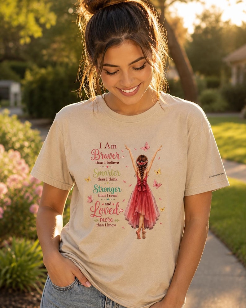 May include: A beige short-sleeved t-shirt featuring a graphic of a person in a red dress with arms raised, surrounded by butterflies. The text reads: I Am Braver than I believe, Smarter than I think, Stronger than I seem, and Loved more than I know.