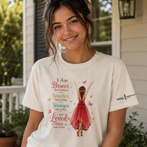 May include: A cream-colored short-sleeve t-shirt featuring a graphic of a person in a red dress with arms raised, surrounded by butterflies. The text reads: I Am Braver than I believe, Smarter than I think, Stronger than I seem and Loved more than I know.